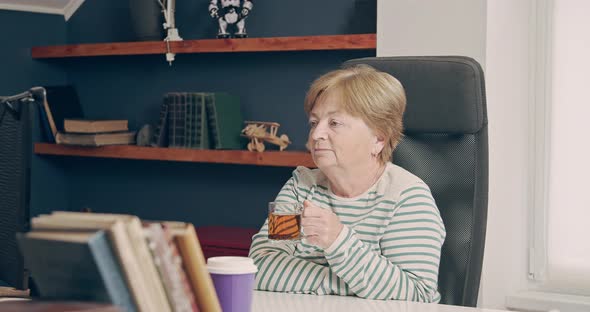 An Elderly Woman Looks Into a Computer Monitor While Working at Home and is About to Drink Tea alt