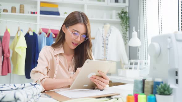 Asian beautiful tailor woman use tablet check new handmade suit order for customer in workshop room. alt