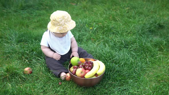 Small Newborn Child in Summer Panama Hat Sit on Grass Barefoot in Bib with Big Bowl of Fresh Fruit