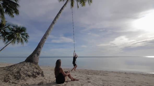 Young caucasian man rope swinging from palm tree on beach with young woman sitting in the sand watch alt