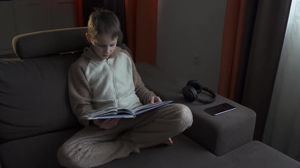 Teen Boy Reads a Book Sitting on the Couch. He Closes the Book and Rests. Quarantine 2020