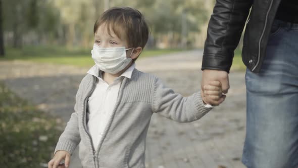 Portrait of Charming Brunette Little Boy in Covid19 Face Mask Walking in Park Holding Hand of alt