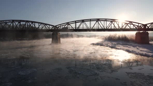 Cinematic Drone Shot Flies Forward Over the Frozen River and Under the Red Steel Bridge at Sunrise