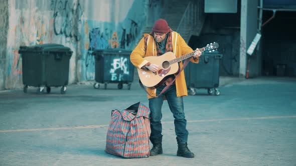 A Poor Man is Playing the Guitar Near Trash Bins alt