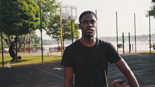 Handsome Afro American Basketball Player is Standing with a Ball on Basketball Court Outdoors alt