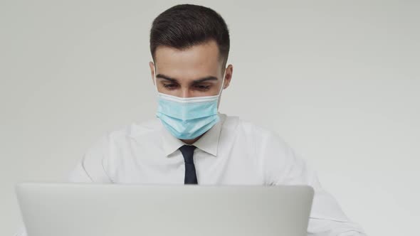 Portrait of Male Student with Medical Mask Thinks During a Studying on a Laptop alt