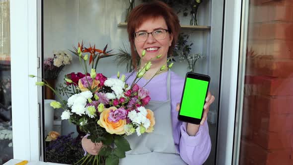 Female florist wearing apron, showing smartphone with chroma key in flower store