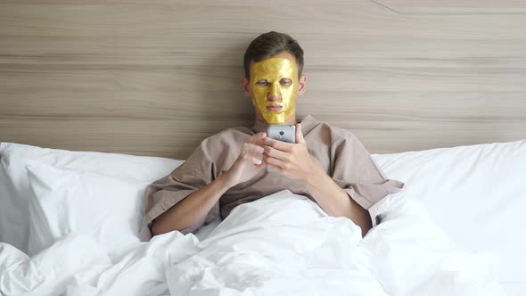 Man with Golden Face Mask Uses Smartphone Lying in Soft Bed alt