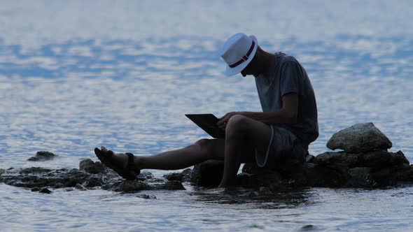 Man Sitting on the Rock on Seaside with Notebook at Dusk. Freelancer Working on the Nature. Hipster