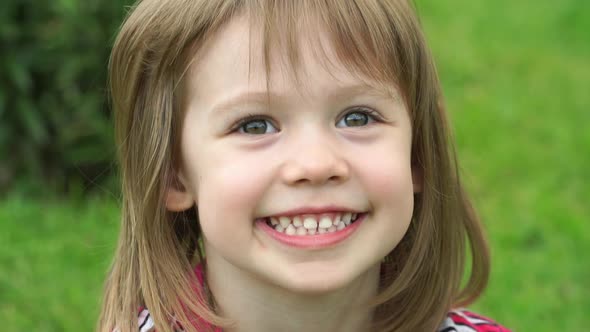 Little Girl in Summer Park Sits on Green Lawn Looks at Camera Smiles and Laughs