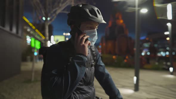 Cyclist in Helmet and Protective Mask Commuter with Bicycle on Way Home From Work Talking on Phone alt