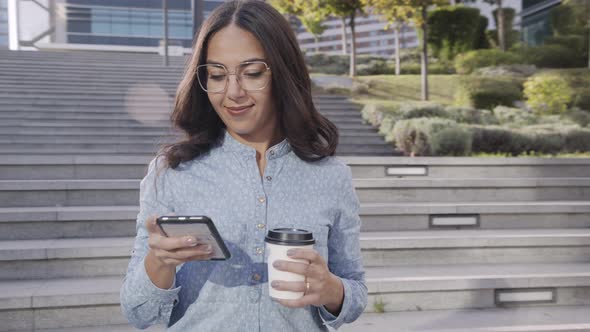 Smiling Hispanic Woman with Coffee To Go Using Smartphone on Stairs alt