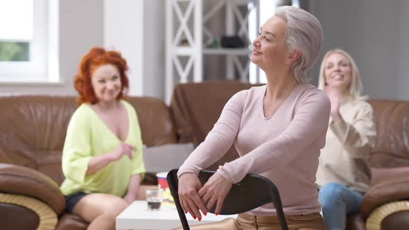 Confident Gorgeous Adult Woman Sitting on Chair at Home Looking Back at Friends Waving and Mocking alt