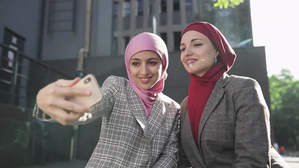 Two Young Muslim Women Wearing Hijab Headscarf Making Selfie at Phone, Sitting Together at Bench