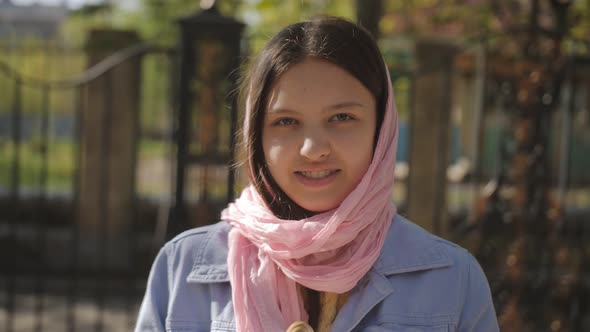 Portrait of a Young Girl with a Pink Scarf on Her Head in the Park Near the Temple alt