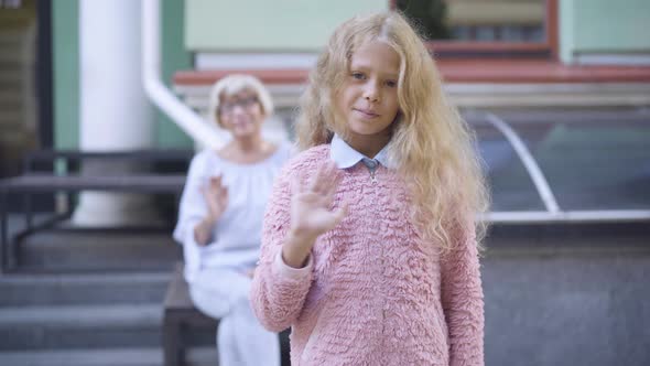 Charming Blond Curly-haired Girl Waving and Smiling at Camera As Blurred Grandmother Sitting at the alt