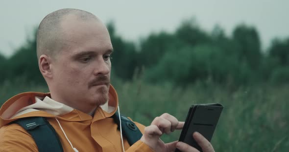Man in a Raincoat is in Nature and Looks at a Map in His Phone