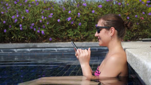 Girl in Black Sunglasses Types on Smartphone Sitting in Pool alt