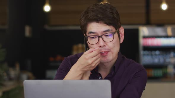Asian man with laptop having a snack and talking on smartphone while sitting at a cafe alt