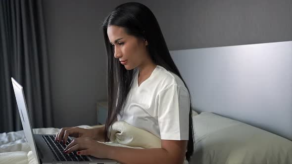 A young woman sitting on a bed in room and works on their laptop.