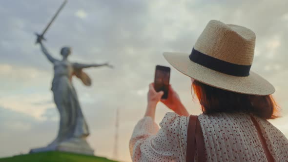 An attractive woman in a hat near Mother Motherland taking a photo on a smartphone on Mamayev Kurgan alt