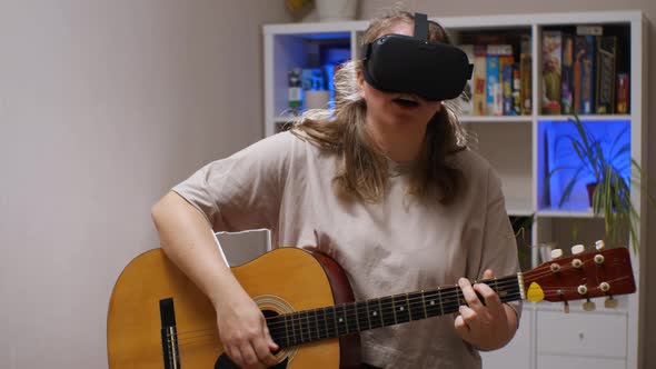 A girl wearing VR glasses sings and plays an acoustic guitar while sitting in a room alt