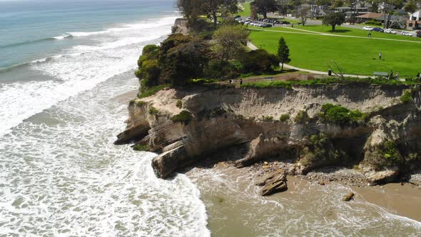 Aerial drone shot over green beach cliffs with grass and trees over the blue ocean waves crashing be alt