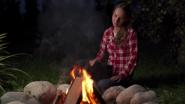 Woman Holds a Cat and Sits Near Campfire at Evening alt