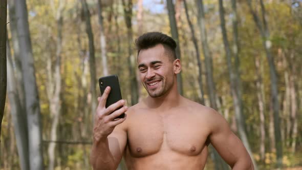 Strong Guy With Naked Torso Stands In Autumn Park And Speaks On Mobile Phone. alt
