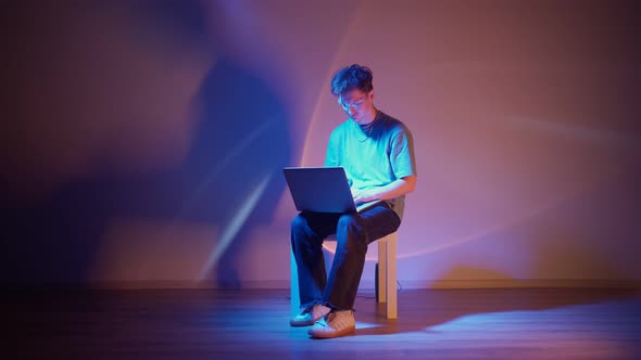 Lonely Stylish Man in Glasses Sits Working at a Laptop in Neon Lighting alt