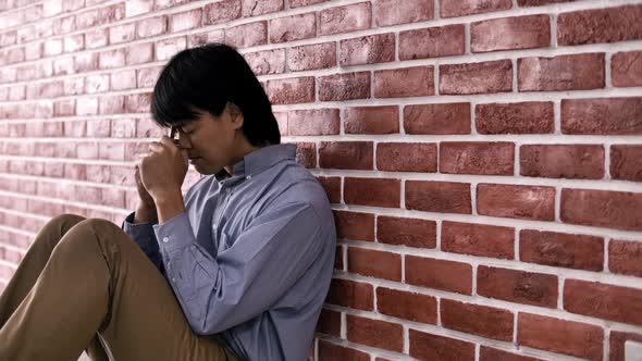 Christian Asian man is sitting against a wall, feeling sad, holding a cross and praying to God. alt