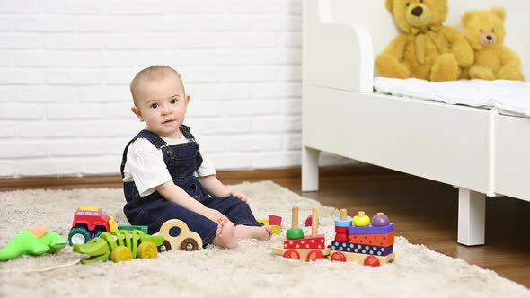 a Baby in a Denim Jumpsuit Sitting Barefoot on the Carpet Smiles and Plays with Toys alt