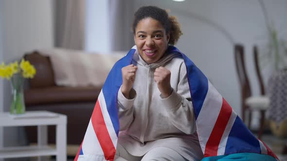 TV POV of Cheerful Smiling African American Woman Wrapped in British Flag Cheering for Soccer Team alt