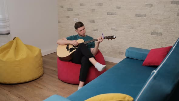 Young Artist Sitting on Red Bean Bag and Playing at the Guitar alt