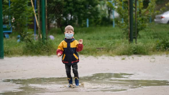 A little boy in a medical mask and rubber boots jumps in puddles and dances