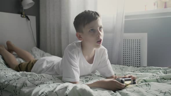 Young Concentrated Pre Teenage Boy Laying on Bed and Playing Video Game Console alt
