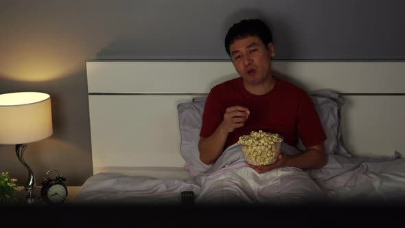 young man watching TV and eating popcorn on a bed at night