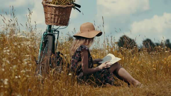 Woman Sitting On Flower Meadow And Reading Book Diary. Tourist Girl Relaxing On Wildflower Field. alt