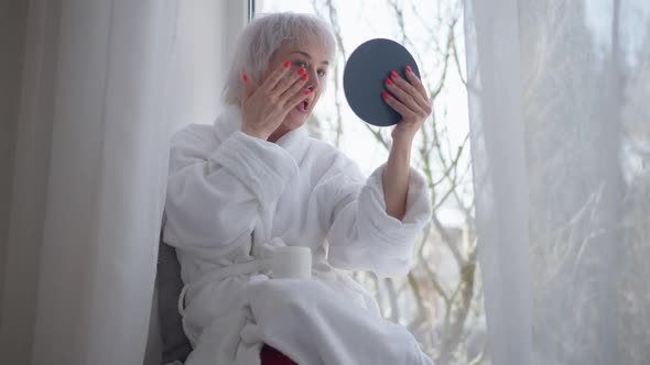 Mature Woman Applying Facial Moisturizer Sitting on Windowsill at Home alt