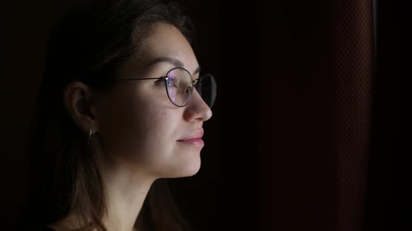 Young woman in glasses looks thoughtfully out the window from a dark room alt