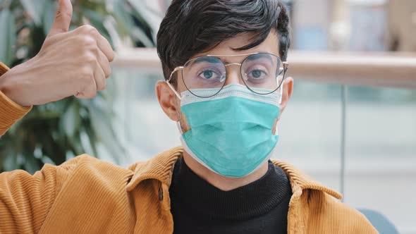 Closeup Young Indian Guy in Medical Mask Posing Indoors Looking at Camera Showing Thumbs Up Sign alt