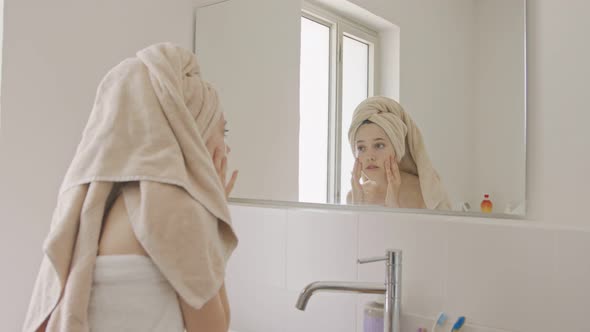 Teenage girl applying moisturizer to her face in front of the bathroom mirror alt
