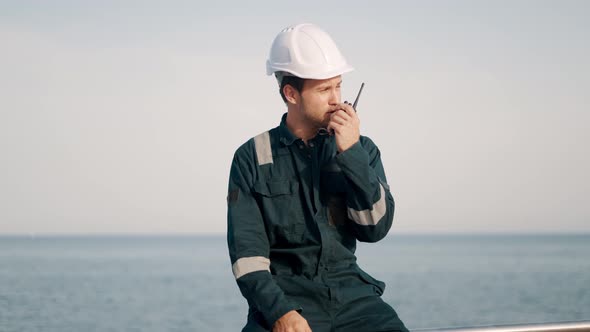 Young Port Worker in Helmet and Coverall Using Vhf Radio Controlling Work Process in Shipping Port alt