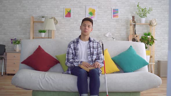 Blind Asian Young Man Reading a Book of Braille Text Sitting on the Couch in the Living Room alt