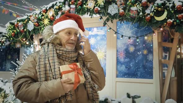 An Elderly Man in a Santa Hat with a Wrapped Gift Box in His Hands is Talking on the Phone at a