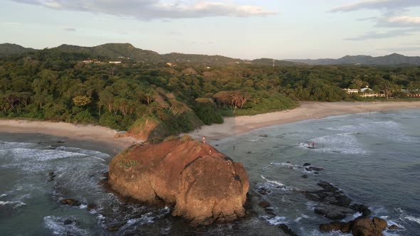 Man standing on the edge of a cliff with his little son. Big rock on a tropical beach with palm tree alt