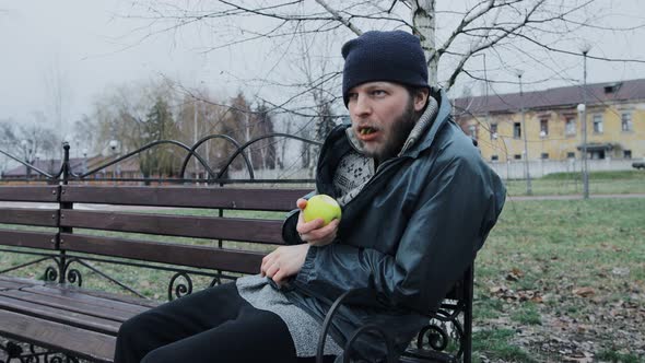 Hungry Bearded Homeless Man Sits on a Bench and Eats a Green Apple in a City Park alt
