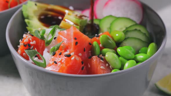 Poke bowl with salmon, rice, avocado, edamame beans, cucumber and radish in gray bowl.  alt