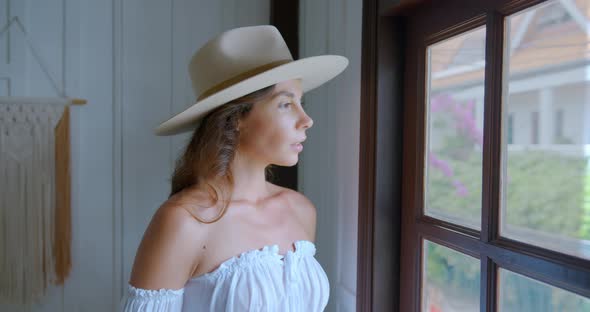 Young Woman Stands By the Wooden Window and Looking in It Her Phone is Ringing and She Answers alt