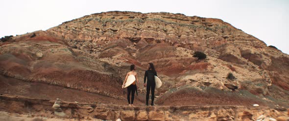 Two surfers standing with their boards and talking in front of a rocky mountain alt